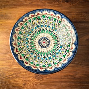 Pode incluir: Uma taça redonda de cerâmica com um padrão radial complexo pintado à mão em tons de verde, azul e vermelho. O design consiste em motivos repetitivos em forma de folha que irradiam de um ponto central, com uma borda festonada azul e branca.