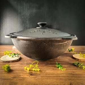 Puede incluir: Una olla de hierro fundido gris oscuro con tapa a juego. La olla tiene dos asas y forma redondeada. La palabra "DRAYALL" está grabada en la tapa. La olla está sobre una superficie de madera con vegetación decorativa.