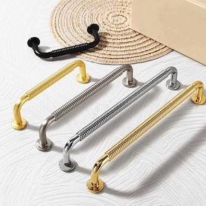 Puede incluir: Un conjunto de tiradores decorativos para armarios en varios acabados metálicos. Los tiradores incluyen opciones doradas, plateadas y negras, cada una con un diseño texturizado único. Los tiradores curvos están dispuestos sobre una superficie clara.