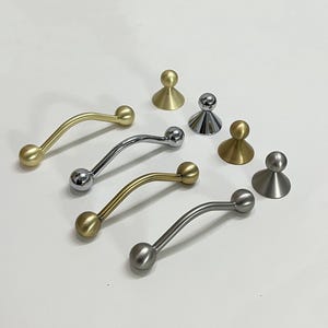 May include: An assortment of metal cabinet hardware, including curved handles and knob pulls. The handles have a curved design with a ball on each end. Finishes include gold, silver, and brushed nickel. The knobs are cone-shaped with a ball on top.