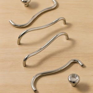 May include: A collection of polished chrome cabinet hardware, including two round knobs and three curved bar pulls. The hardware is arranged on a light wood surface, showcasing the reflective quality of the metal.