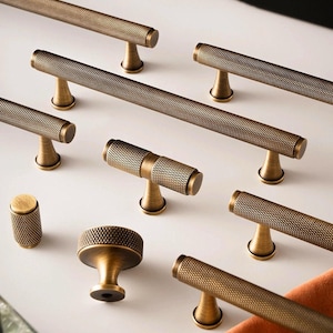 May include: A collection of antique brass cabinet hardware, including bar pulls and knobs. The hardware features a textured, diamond-patterned design. The pulls are various lengths, and the knobs are round with a similar texture. The hardware is displayed on a white surface.