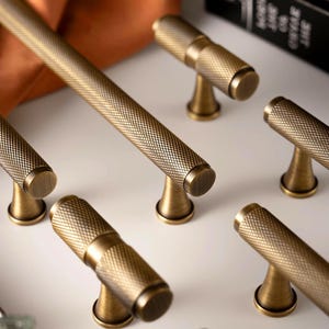 May include: Close-up of several antique brass cabinet pulls and a long handle. The hardware features a textured, diamond-patterned design. The pulls have a T-shape, while the handle is a long bar. The hardware is on a white surface.