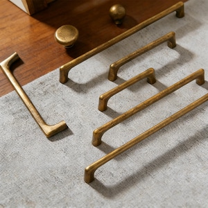 Pode incluir: Uma coleção de ferragens de armário em latão antigo, incluindo várias puxadores de barra e um botão redondo. O hardware é disposto sobre uma superfície cinza claro, mostrando o acabamento envelhecido e o design. Os puxadores variam em comprimento.