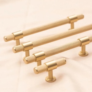 May include: Four gold-toned cabinet pulls with a textured, diamond-patterned grip. The pulls are cylindrical with a circular base and are arranged at different lengths. The hardware is set against a soft, neutral background.