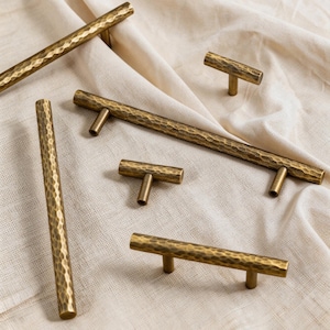 Puede incluir: Un conjunto de tiradores y pomos de armario de latón martillado. La ferretería tiene una superficie texturizada y un tono dorado cálido. Los tiradores son de varios tamaños y formas, incluyendo tiradores de barra y pomos en forma de T, dispuestos sobre una tela color crema.