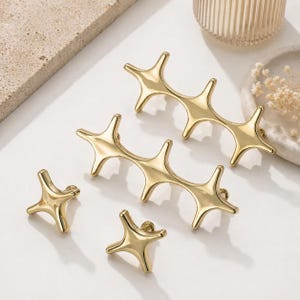 May include: A collection of gold-colored cabinet hardware, including star-shaped knobs and elongated pulls. The hardware has a modern, minimalist design, with a polished finish. The arrangement suggests a focus on home decor and interior design.