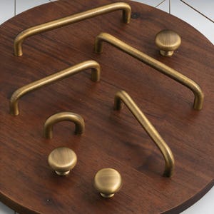 Puede incluir: Colección de herrajes de latón antiguo para armarios, que incluye tiradores de barra y pomos redondos de varios tamaños. Los herrajes se exhiben sobre una superficie de madera marrón oscuro, mostrando los tonos cálidos del metal.