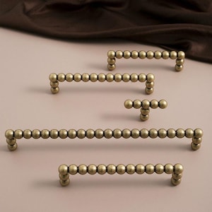 May include: Several antique brass cabinet pulls in various sizes are arranged on a neutral surface. Each pull is composed of small, evenly spaced spheres, creating a beaded appearance. The hardware is designed for furniture and adds a vintage touch.