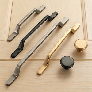 Puede incluir: Conjunto de herrajes para armarios en varios acabados metálicos. Incluye tiradores plateados, negros y dorados, junto con pomos a juego. La ferretería se muestra sobre una superficie de madera clara.