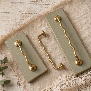 Puede incluir: Tres tiradores ondulados de latón sobre tablas rectangulares de madera gris verdoso claro. Los tiradores tienen una forma orgánica única y están montados con herrajes redondos de color latón. Las tablas descansan sobre una tela de tono neutro.