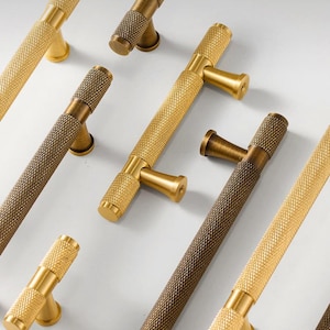 Pode incluir: Uma variedade de puxadores de armário dourados e bronzeados. Os puxadores têm uma pega texturizada com padrão de diamante e um design em forma de T. O hardware está disposto sobre um fundo claro, mostrando os diferentes acabamentos e estilos. Medem cerca de 15 cm.