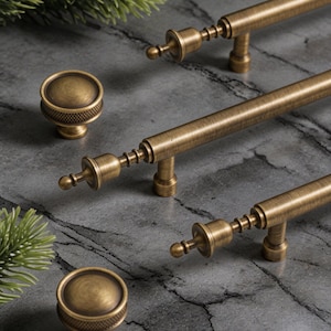 May include: Antique brass cabinet hardware set, including two bar pulls and two round knobs. The pulls have a detailed design with a central bar and decorative ends. The knobs feature a textured surface. The hardware is displayed on a gray marble surface.
