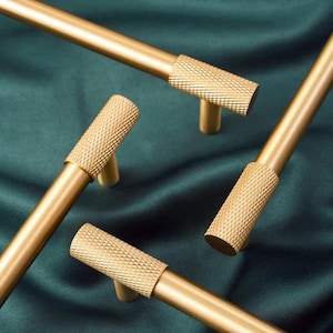 Op de afbeelding: Close-up van verschillende goudkleurige kastgrepen met een getextureerde, diamantpatroon grip. De grepen staan tegen een donkergroene stoffen achtergrond, waardoor de metallic afwerking wordt benadrukt. Ideaal voor keuken- of badkamerkasten.