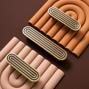 May include: Three antique brass cabinet pulls with a grooved design are displayed on a brown background. The pulls have a rectangular shape with rounded ends and a series of parallel grooves. The background features terracotta-colored, arched shapes.