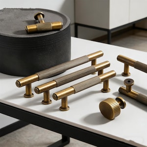 May include: A collection of antique brass cabinet hardware, including T-bar handles and a round knob. The handles feature a ribbed design on the bar and are displayed on a white surface. The hardware is designed for furniture and home decor.