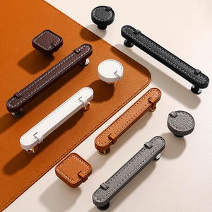 May include: A collection of cabinet hardware, including knobs and pulls, in various colors. The hardware features a leather-like texture with contrasting stitching. Colors include brown, white, gray, and black. The pulls are approximately 6 inches long.