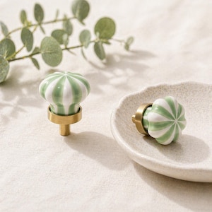 May include: Two ceramic cabinet knobs with a green and white striped design. The knobs have a rounded shape and are mounted on brass hardware. One knob is upright, and the other is lying on a decorative plate.