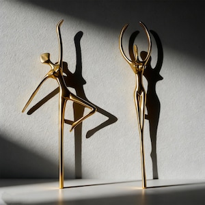 Peut inclure: Deux sculptures dorées en métal représentant des danseurs. L'un des danseurs est en pose avec un bras tendu et une jambe pliée, tandis que l'autre a les bras levés. Les sculptures projettent de longues ombres sur le fond blanc.