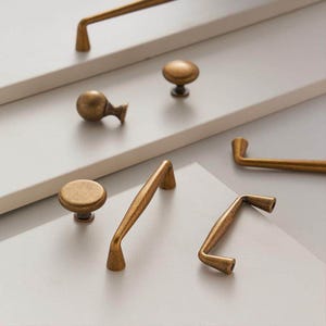 Pode incluir: Uma variedade de ferragens de armário em latão antigo, incluindo puxadores e puxadores. As ferragens apresentam um acabamento quente e envelhecido e vários designs, como puxadores redondos, puxadores curvos e puxadores de barra. As ferragens são exibidas em uma superfície cinza claro neutra.