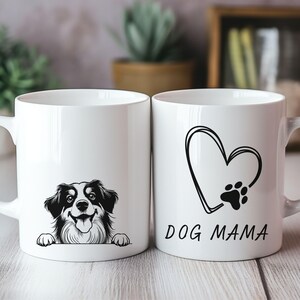 Peut inclure: Deux mugs en c&eacute;ramique blancs. L'un pr&eacute;sente une illustration en noir et blanc d'un chien regardant par-dessus le bord. L'autre mug a un c&oelig;ur avec une empreinte de patte et les mots "DOG MAMA" en noir.