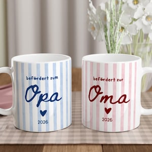 Könnte beinhalten: Zwei weiße Keramiktassen mit vertikalen Streifen. Die linke Tasse hat blaue Streifen, den Text "befördert zum Opa", ein blaues Herz und "2026". Die rechte Tasse hat rosa Streifen, den Text "befördert zur Oma", ein rotes Herz und "2026".