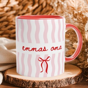 Könnte beinhalten: Eine Keramiktasse mit rotem Innenraum und Henkel, die außen weiß mit vertikalen, hellrosa Wellenstreifen gestaltet ist. Der Text "emmas oma" ist in roter Schreibschrift über einer roten Schleifengrafik auf der Vorderseite gedruckt.