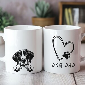 Peut inclure: Deux mugs en c&eacute;ramique blancs. L'un pr&eacute;sente une illustration de chien en noir et blanc. L'autre mug a un c&oelig;ur et une empreinte de patte avec le texte "DOG DAD" en noir. Les deux mugs ont une anse blanche.