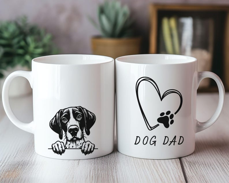 Peut inclure: Deux mugs en c&eacute;ramique blancs. L'un pr&eacute;sente une illustration de chien en noir et blanc. L'autre mug a un c&oelig;ur et une empreinte de patte avec le texte "DOG DAD" en noir. Les deux mugs ont une anse blanche.
