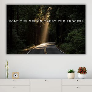 May include: A rectangular wall art print featuring a dark forest road with a bright beam of light shining through the trees. The text "HOLD THE VISION, TRUST THE PROCESS" is printed in white capital letters across the centre of the image.