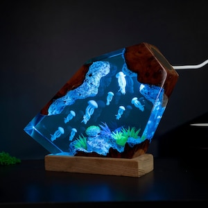 May include: A unique, illuminated resin lamp featuring a marine scene with jellyfish. The lamp has a wooden base and a clear resin body with embedded jellyfish and coral. The lamp's interior glows with a blue light, creating a captivating underwater effect.