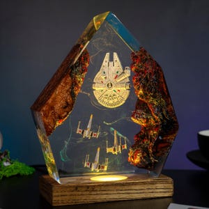 Peut inclure: Une œuvre d'art en résine illuminée représentant le Faucon Millenium et des chasseurs X-wing. L'œuvre est enfermée dans un bloc de résine clair et géométrique, posé sur une base en bois. La pièce mesure environ 20 cm de haut.