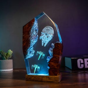 May include: A Star Wars-themed lamp featuring the Millennium Falcon and X-wing fighters encased in clear resin. The lamp has a wooden base and a blue LED light, with wood and blue accents.