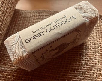 May include: A rectangular bar of soap wrapped in a beige net and paper label. The label reads "jewelweed infused GREAT OUTDOORS" with a small illustration. The soap rests on a burlap surface.