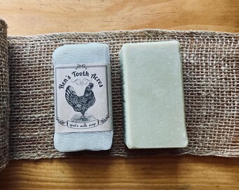 May include: Two rectangular bars of soap. One bar is wrapped in a light blue mesh fabric with a paper label featuring a black illustration of a hen and the text Hen's Tooth Acres goat's milk soap. The second bar is solid pale green.