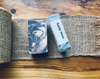May include: Two bars of soap on a burlap ribbon. One bar has a marbled pattern in shades of gray and cream. The other bar is wrapped in a light blue mesh with a paper label that reads "Earthy Sweet."