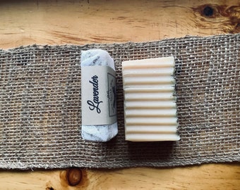 May include: A white bar of soap with a ribbed texture, wrapped in a white mesh bag with a brown label that says "Lavender". The soap and bag are on a brown burlap surface.