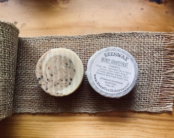 May include: Two round soaps on a burlap ribbon. One soap is light brown with dark specks. The other soap is wrapped in clear plastic with a label that reads "BEESWAX BERRY GRAPEFRUIT" and lists ingredients. The background is a wooden surface.
