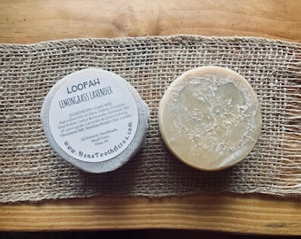 May include: Two round soaps on a burlap cloth. One soap is wrapped with a label that reads "LOOFAH LEMONGRASS LAVENDER" and lists ingredients. The other soap is a light beige color with a textured surface.