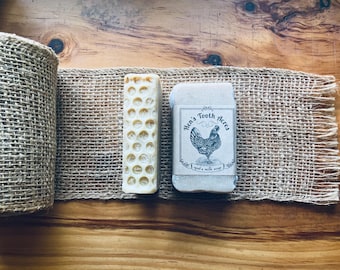May include: A rustic display featuring a roll of burlap, a bar of soap with a honeycomb pattern, and a wrapped soap bar labeled "Hen's Tooth Acres" with a chicken illustration. The items are arranged on a wooden surface.
