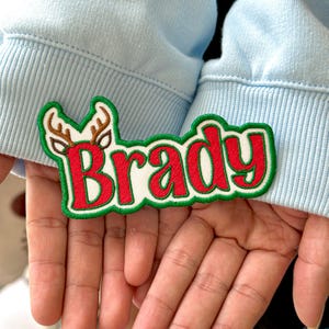 May include: Embroidered patch with the name "Brady" in red letters with a white outline, set against a green border. A brown reindeer head with antlers is on the left side. The patch is held in front of a light blue sweatshirt.