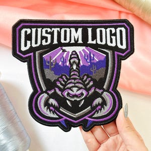 May include: A shield-shaped embroidered patch featuring a central scorpion graphic in shades of purple, grey, and black. The background depicts a desert landscape with cacti and a sunburst. The text CUSTOM LOGO is stitched in white at the top.