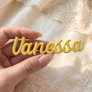 May include: A yellow embroidered patch with the name "Vanessa" in a cursive font. The patch is held in a person's hand against a soft, off-white background. The thread has a slight sheen.