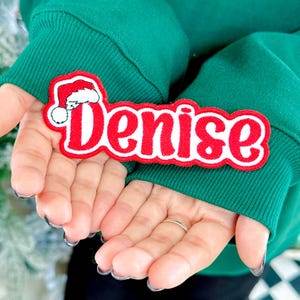 May include: A red and white embroidered patch with the name "Denise" in a festive font, topped with a Santa hat. The patch is held in open hands, against a green background.