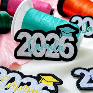 May include: Embroidered graduation patches with the year 2026 and the name "Camila" in a script font. The patches have a black border and a graduation cap design. Spools of colorful thread are in the background.