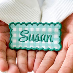 May include: A rectangular embroidered patch with a scalloped edge featuring a light green and white gingham pattern. The name Susan is stitched in the center in a dark green cursive font. The patch has a dark green border.