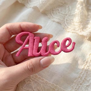 May include: A pink embroidered patch with the name "Alice" in a cursive font. The patch is held in a hand against a background of white lace fabric. The letters are a vibrant, saturated pink.