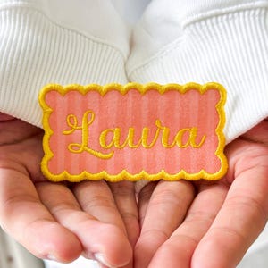 May include: A rectangular patch with a scalloped edge featuring a light coral and white vertical striped pattern. The name Laura is embroidered in the center in a yellow cursive script font. The patch is positioned against white fabric.