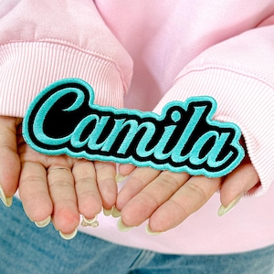 May include: A teal and black embroidered patch with the name "Camila" in a cursive font. The patch is held in open hands, against a light pink background. The patch has a black base with teal stitching.