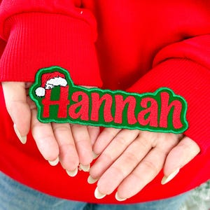 May include: A red and green embroidered patch with the name "Hannah" in red script. A small Santa hat is embroidered above the name. The patch is held against a red background.